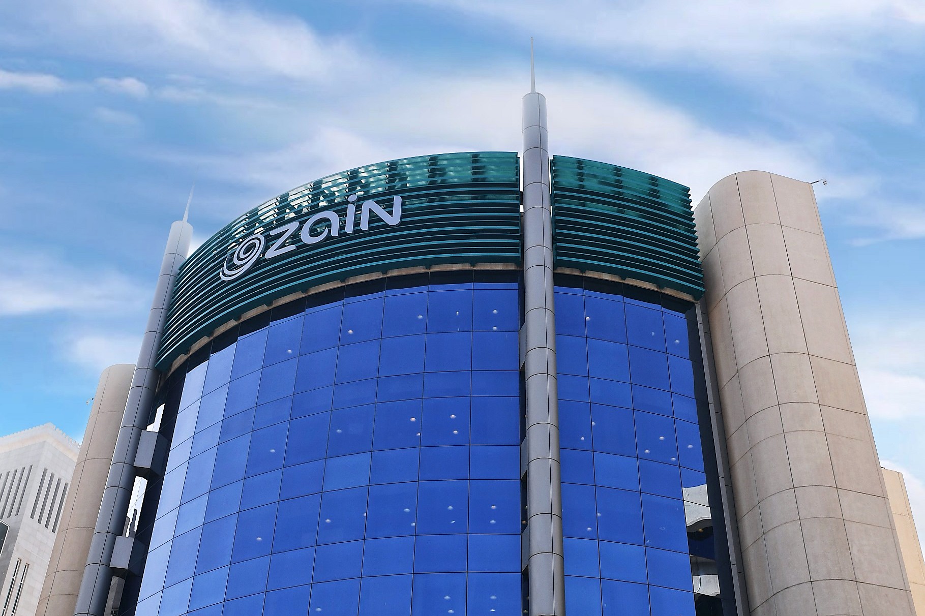 The Zain headquarters tower in Kuwait, a curved facade of blue glass with the Zain logo across its upper band.